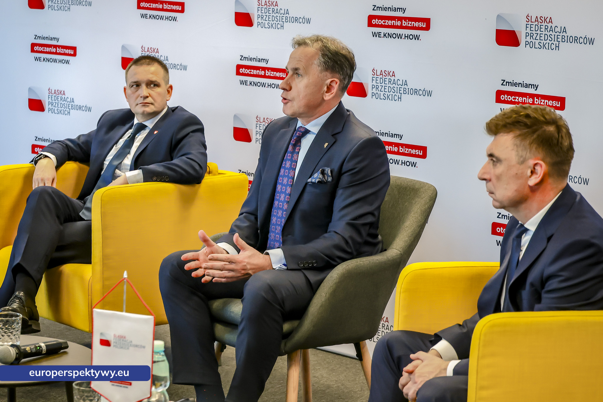 Trzech panelistów w żółtych i szarych fotelach podczas dyskusji