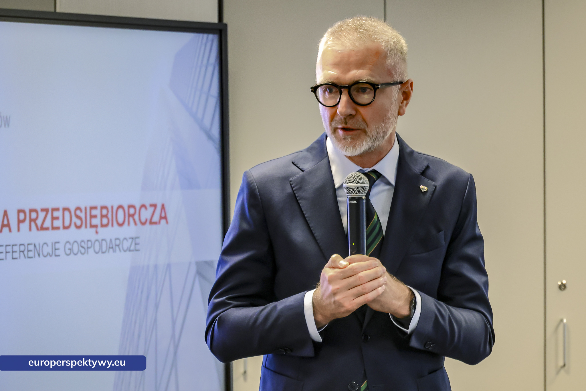 Uczestnik przemawia z mikrofonem przed ekranem z nazwą konferencji
